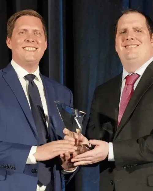 Two men in suits smiling while presenting a glass award at a formal ceremony