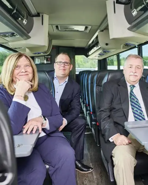 Three professionally dressed passengers seated comfortably inside a modern charter bus with overhead storage and fold-down tray tables