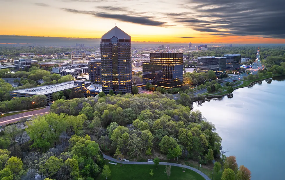 Aerial view of a suburban city skyline at sunset, with glass office towers, tree-lined parks, and a calm lake in the foreground