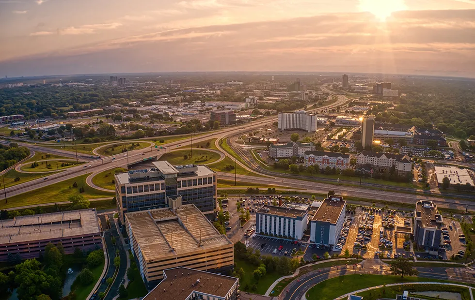 Aerial view of a Midwest city at sunset, showing office buildings, highway interchanges, and urban sprawl extending to the horizon