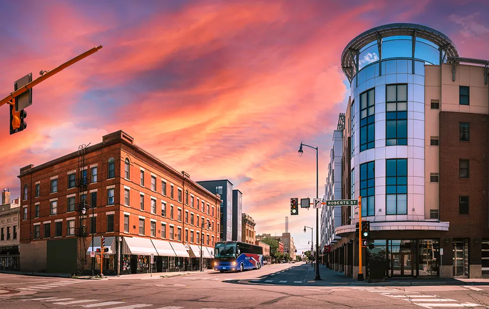 A charter bus travels through a downtown intersection at sunset, passing historic brick buildings and a modern glass-facade building on Roberts St
