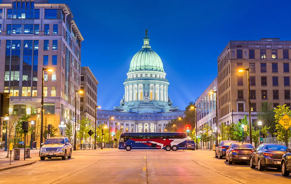 Lorenz Bus Service coach parked on a downtown Madison street in front of the illuminated Wisconsin State Capitol at dusk