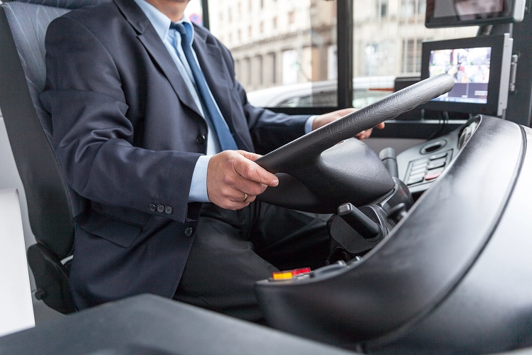 Professional bus driver in a suit at the wheel, with dashboard controls and a monitor visible in the cab