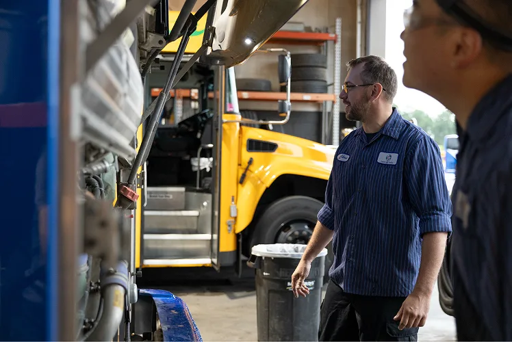 Lorenz technician inspecting a bus in the maintenance garage with a yellow school bus visible in the background