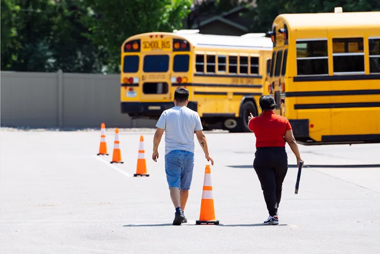 Two school bus drivers walking through a cone-marked training course toward parked yellow school buses
